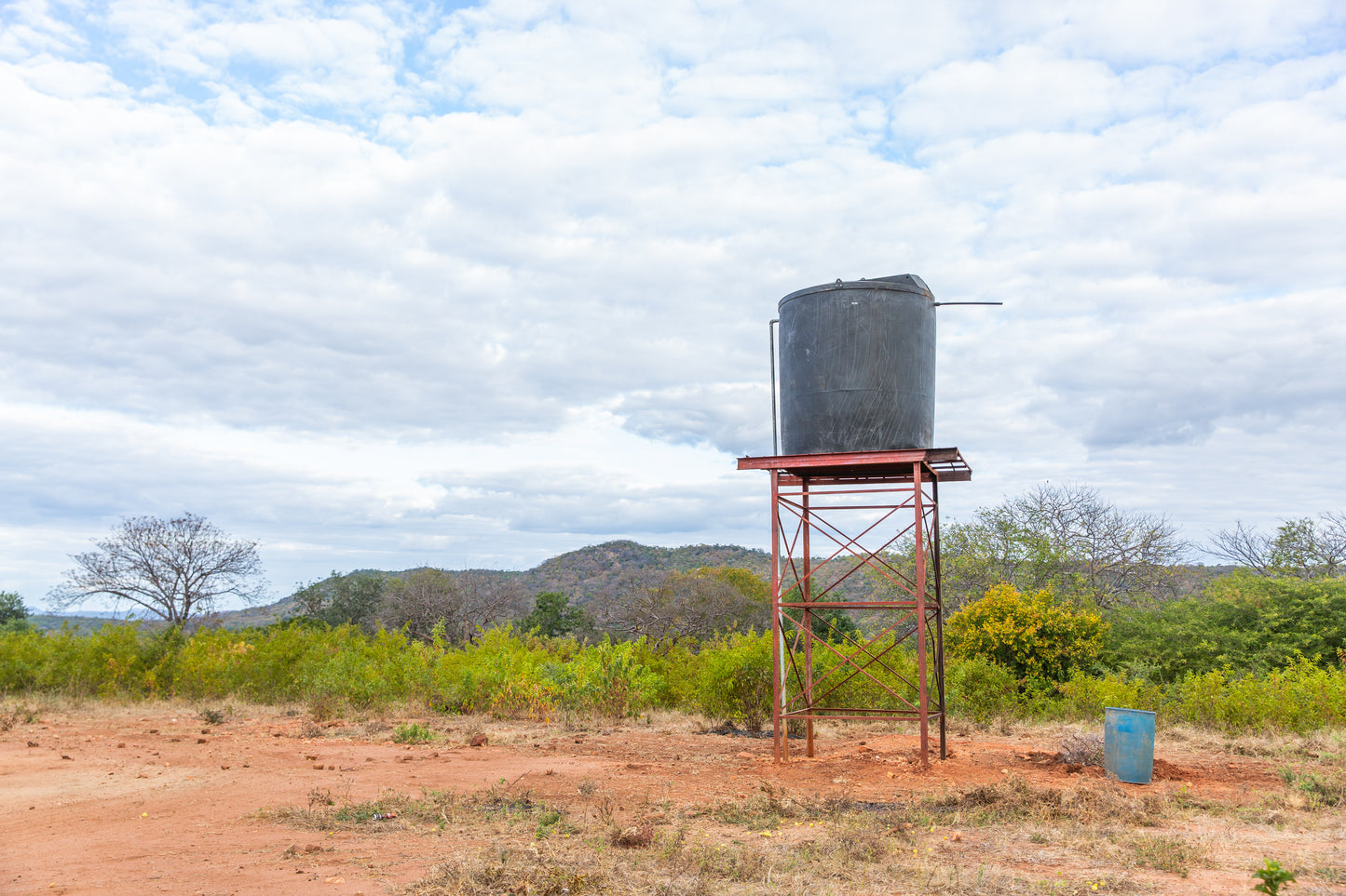 A large black water tank on a red metal elevated stand in an outdoor rural setting with reddish-brown ground, green shrubs, distant hills, and a cloudy blue sky.