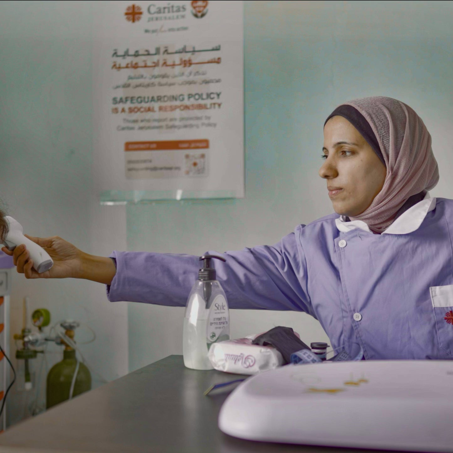 A woman in a purple uniform and mauve hijab stands behind a counter, holding a white device. A hand sanitizer bottle and other items sit on the counter, with a Caritas banner visible on the wall behind her.