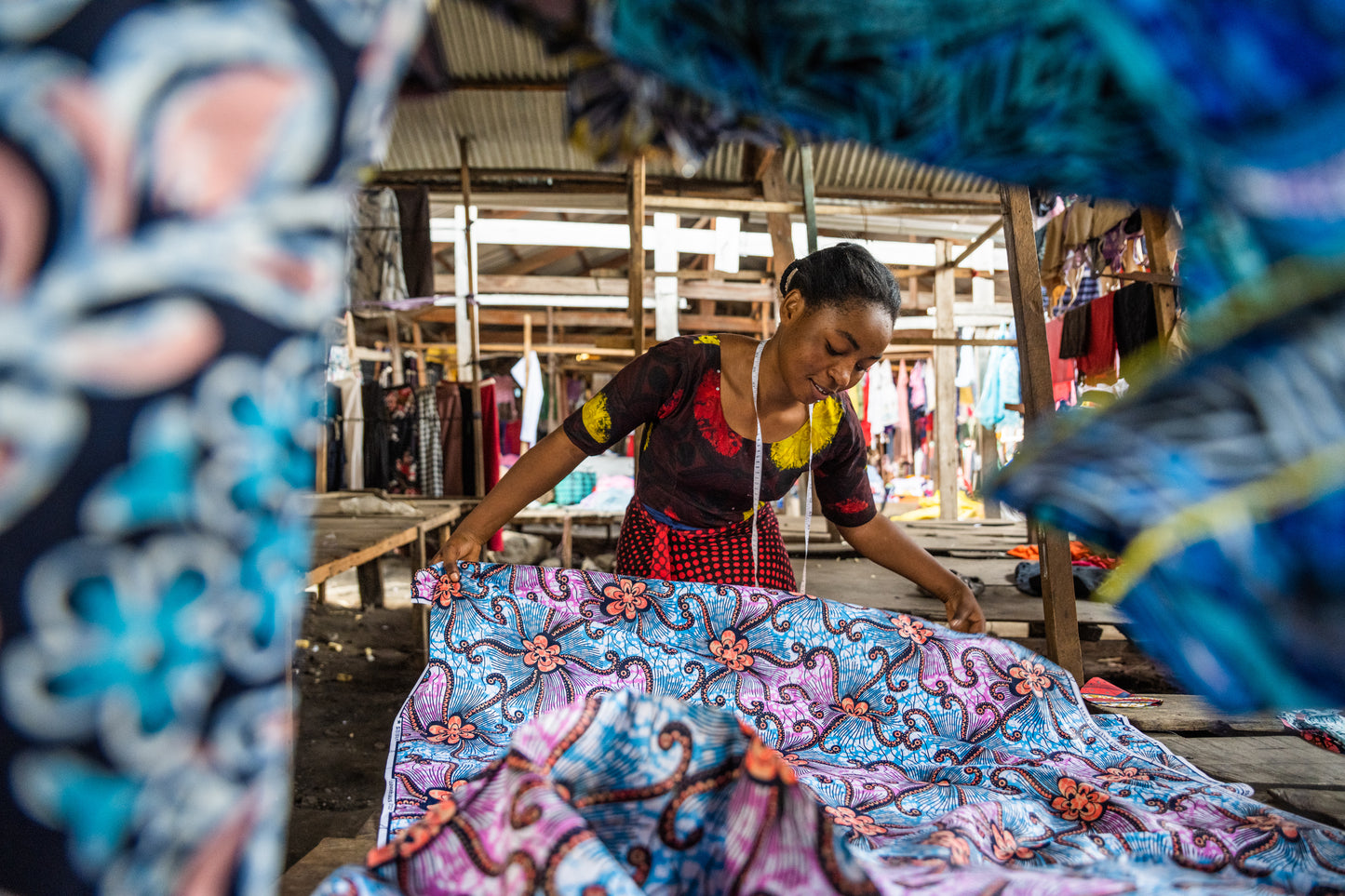 A woman with a tape measure around her neck laying out colourful patterned fabric at an outdoor stall, surrounded by vibrant textiles