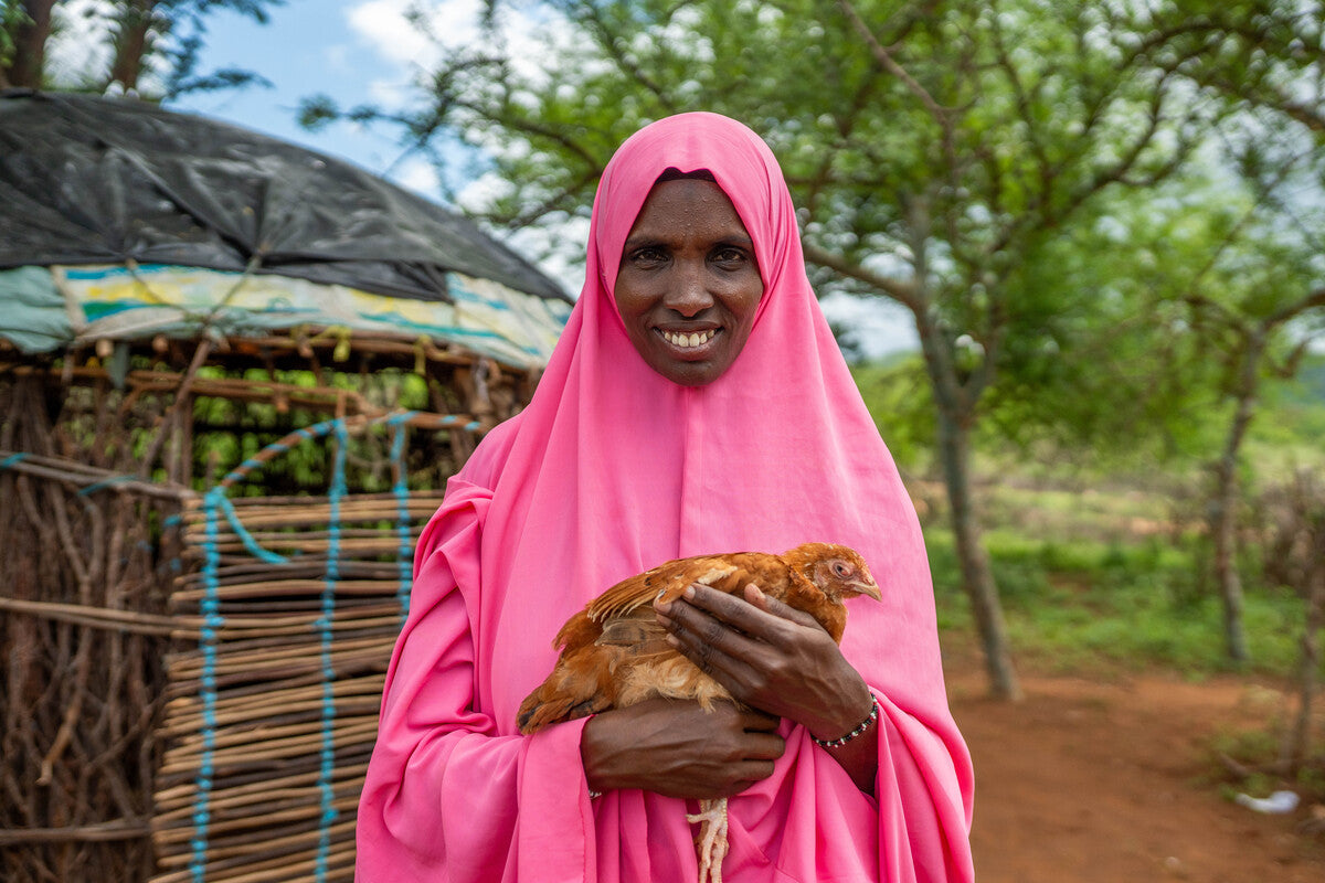 A smiling woman wearing a bright pink hijab holding a brown hen, standing outside a rural homestead surrounded by trees