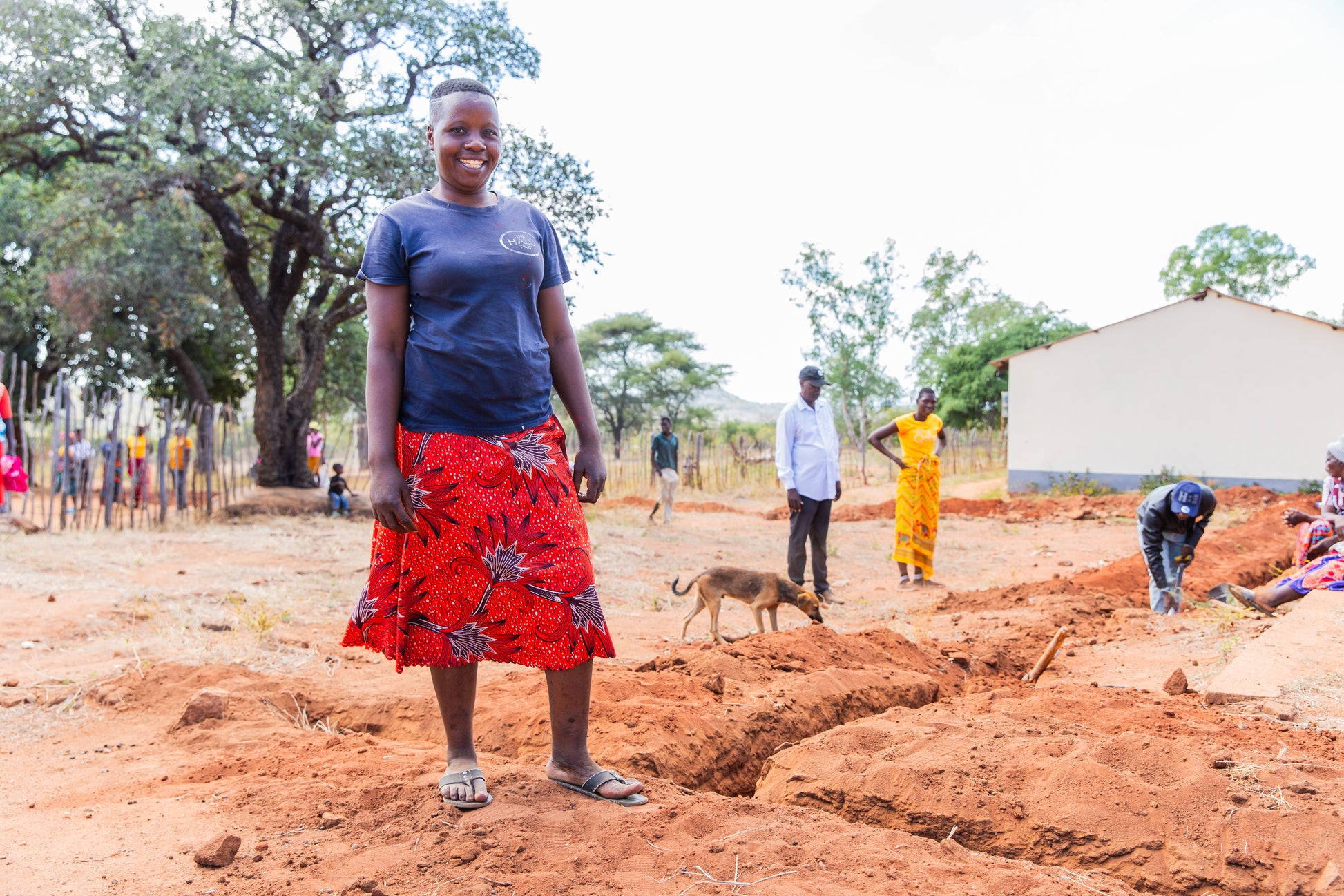 A woman smiles at a community water supply construction site in rural Africa, with villagers working in the background.