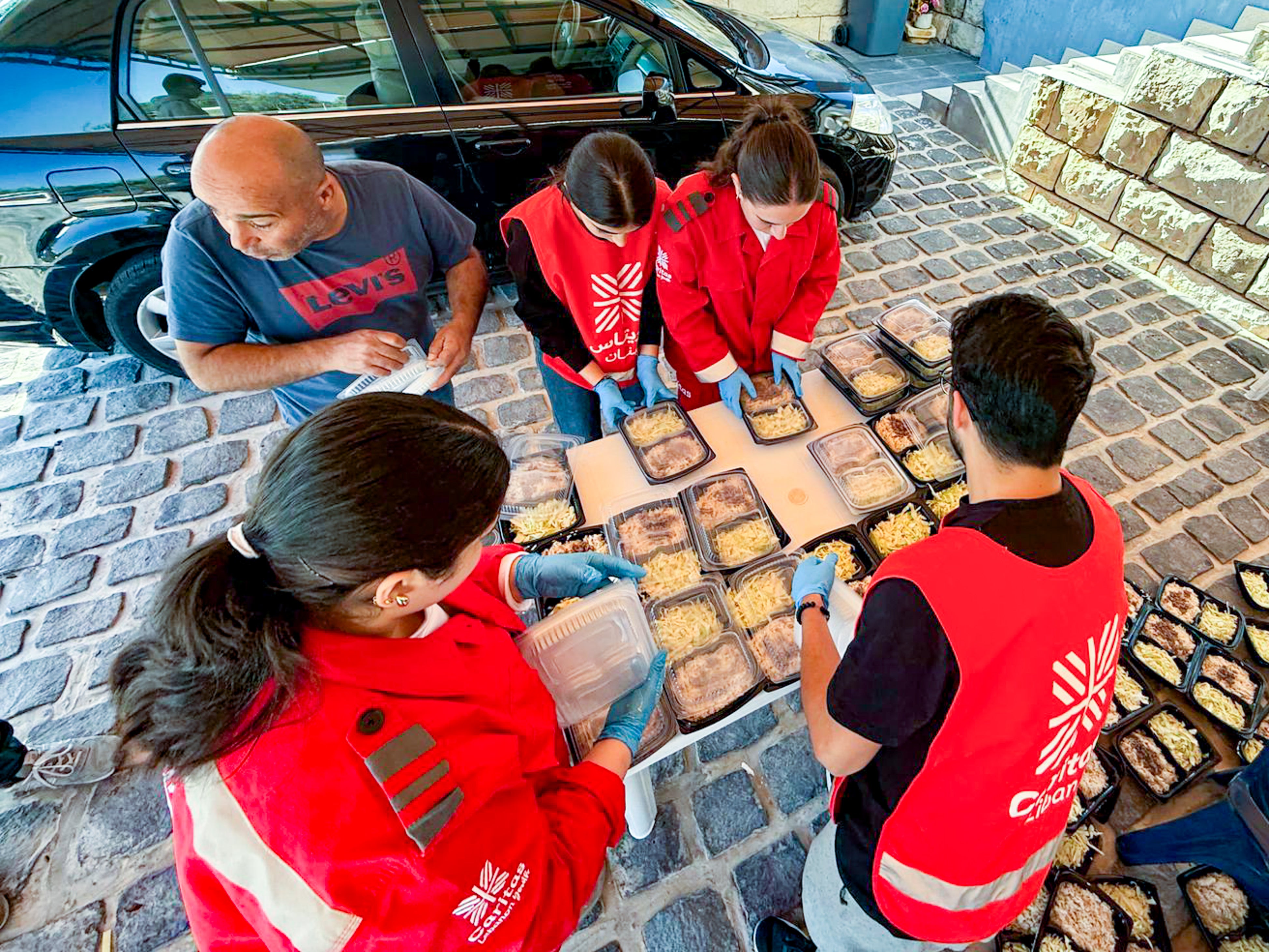 Several people in red service vests and blue gloves are preparing food trays outdoors on a cobblestone surface next to a black car. The scene shows a collaborative effort, with one person holding a clear container and others arranging packaged meals.