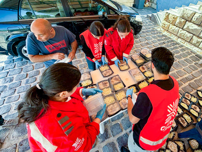 Several people in red service vests and blue gloves are preparing food trays outdoors on a cobblestone surface next to a black car. The scene shows a collaborative effort, with one person holding a clear container and others arranging packaged meals.