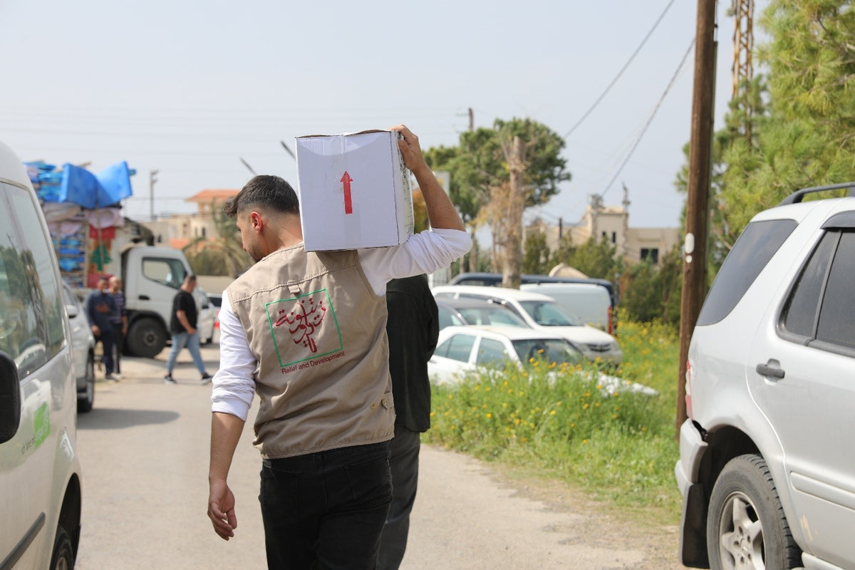 A man in a branded tabard carrying a large white relief box on their shoulder along a road, with vehicles and other workers visible in the background