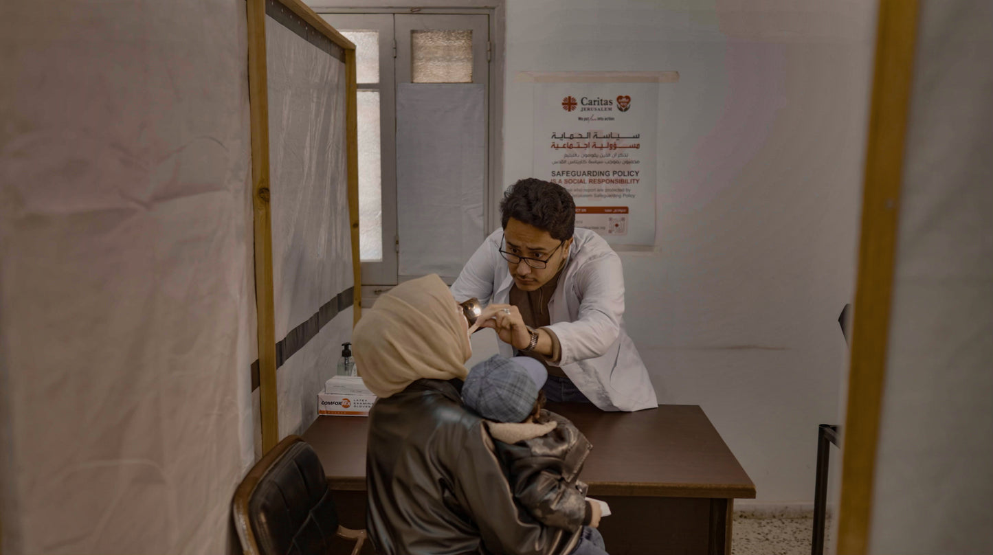 A doctor in a white lab coat examines a person wearing a beige headscarf and a blue face mask inside a clinical examination room with a desk and medical posters on the wall.