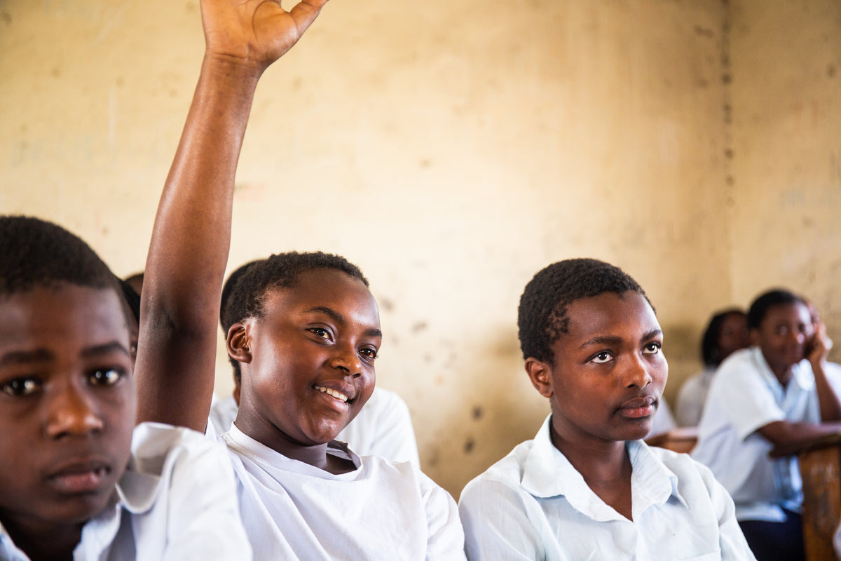A smiling child in a white school uniform raises their hand in a classroom, with other children in white uniforms seated at desks in the background.