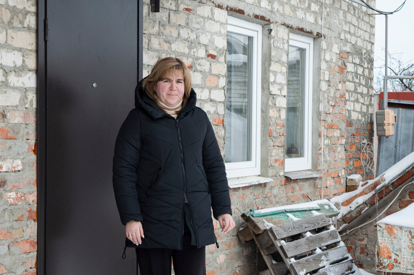 Woman wearing a black coat standing in front of a brick wall with a window.