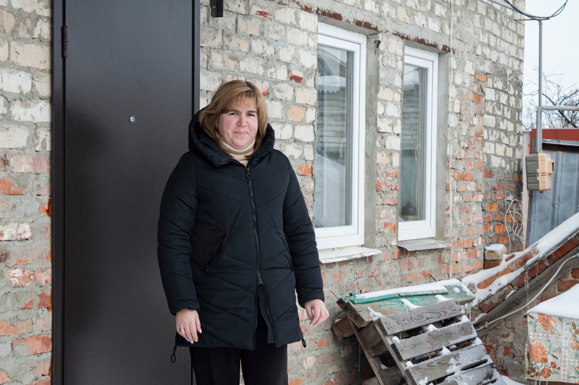 Woman wearing a black coat standing in front of a brick wall with a window.