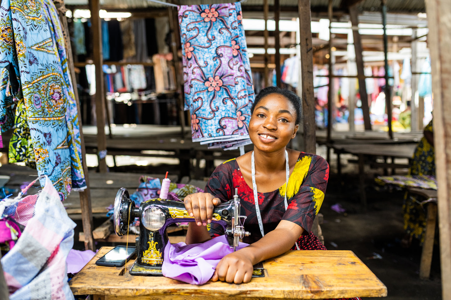 A smiling woman with a tape measure around her neck using a sewing machine at an outdoor stall, surrounded by colourful patterned fabrics