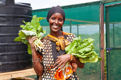 A smiling woman wearing a black headwrap and a black, orange, and white patterned dress holds two large bunches of fresh green leafy vegetables in front of a greenhouse with a black storage tank.