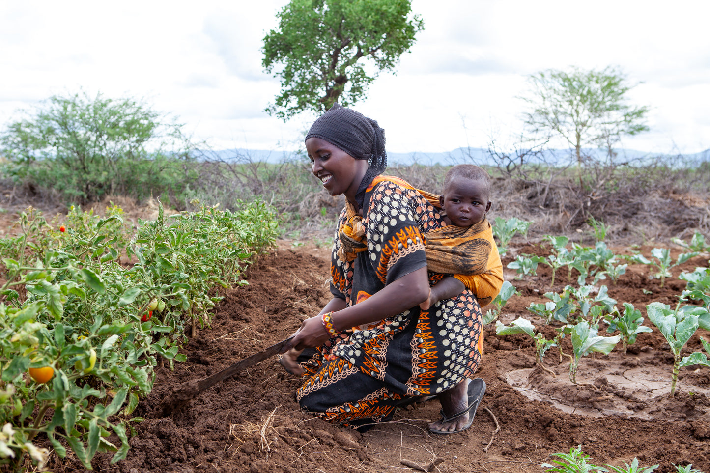 A smiling woman in a patterned dress tending to a vegetable garden with a hand tool, with a young baby carried on her back, in a rural landscape