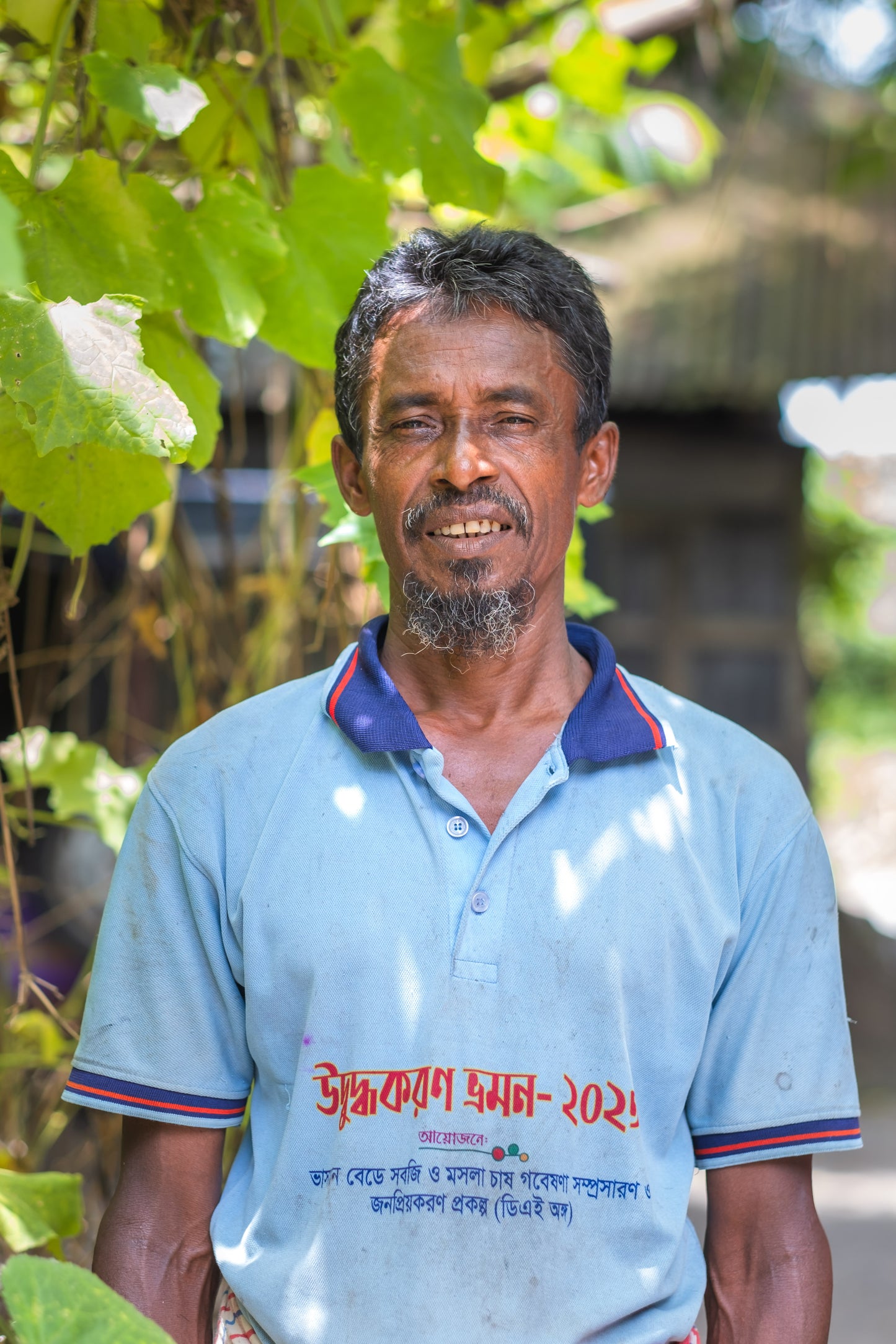 A smiling man wearing a light blue polo shirt with red and navy trim, standing outdoors under green leaves. The shirt has red and blue text printed on the front in Bengali script.